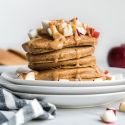 Apple cinnamon pancakes stacked on a plate with melted peanut butter and chopped apples.