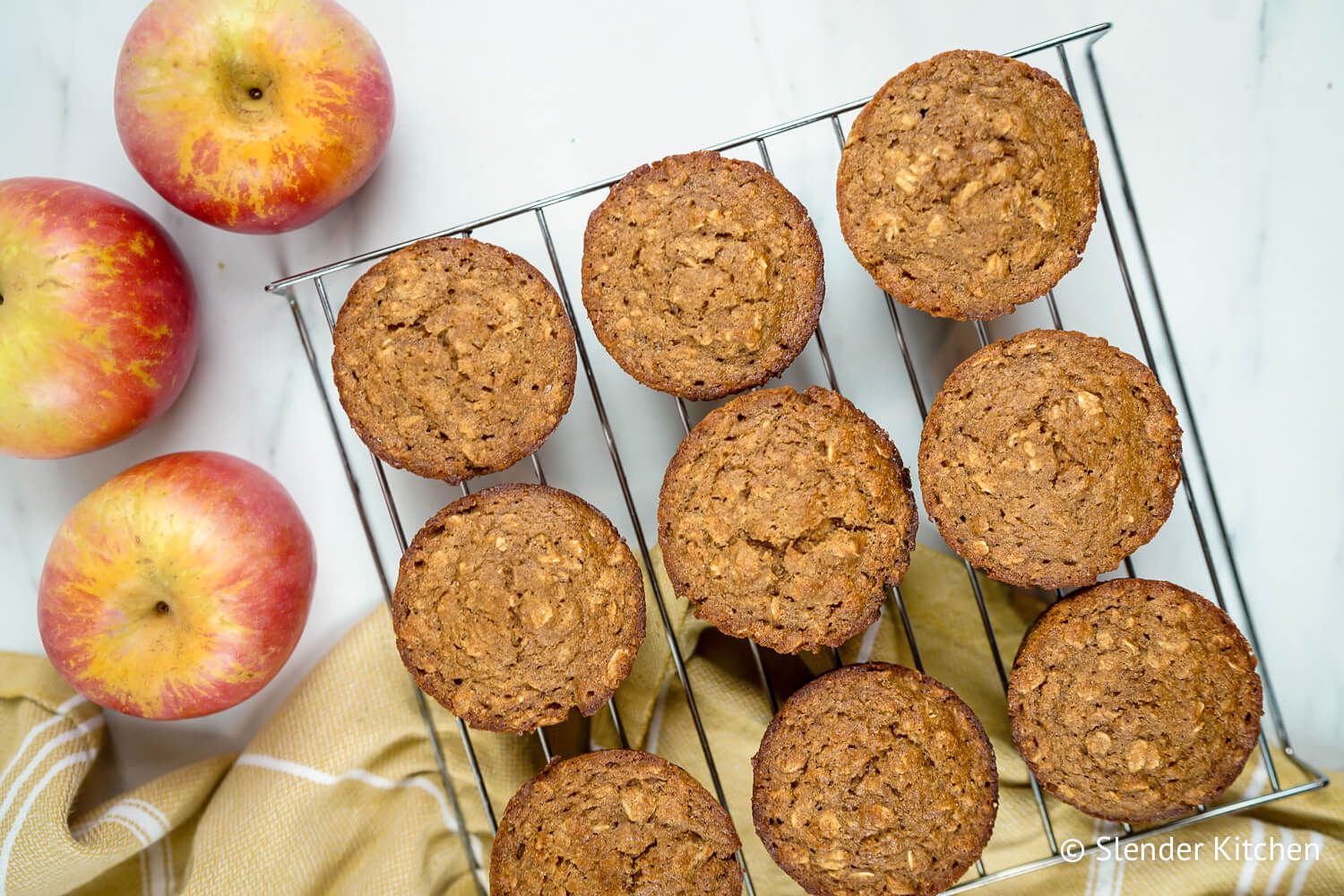 Applesauce oatmeal muffins with oatmeal flecks on a baking rack with a yellow napkin and apples.