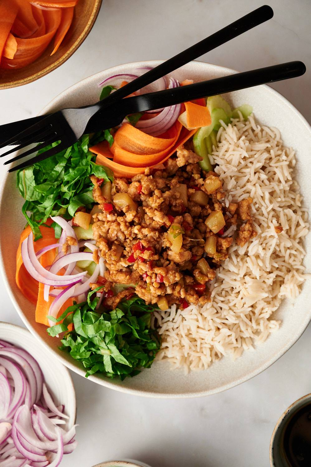 Asian ground turkey and rice bowls with carrots, lettuce, and onions in a white bowl with silverware.