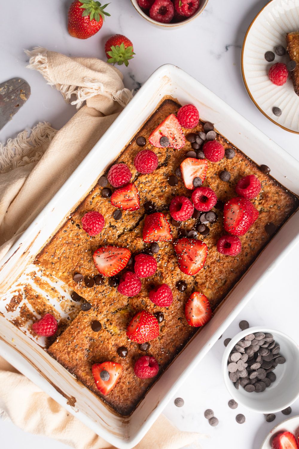 Baked rolled oats with bananas, old fashioned oats, and almond milk in a baking dish with berries and chocolate chips.