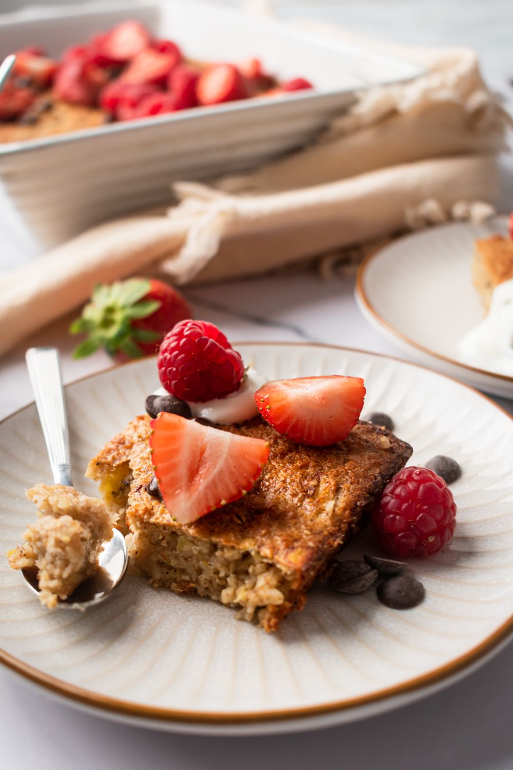 Slices of baked oatmeal on a plate with a spoon, sliced berries, and chocolate chips.