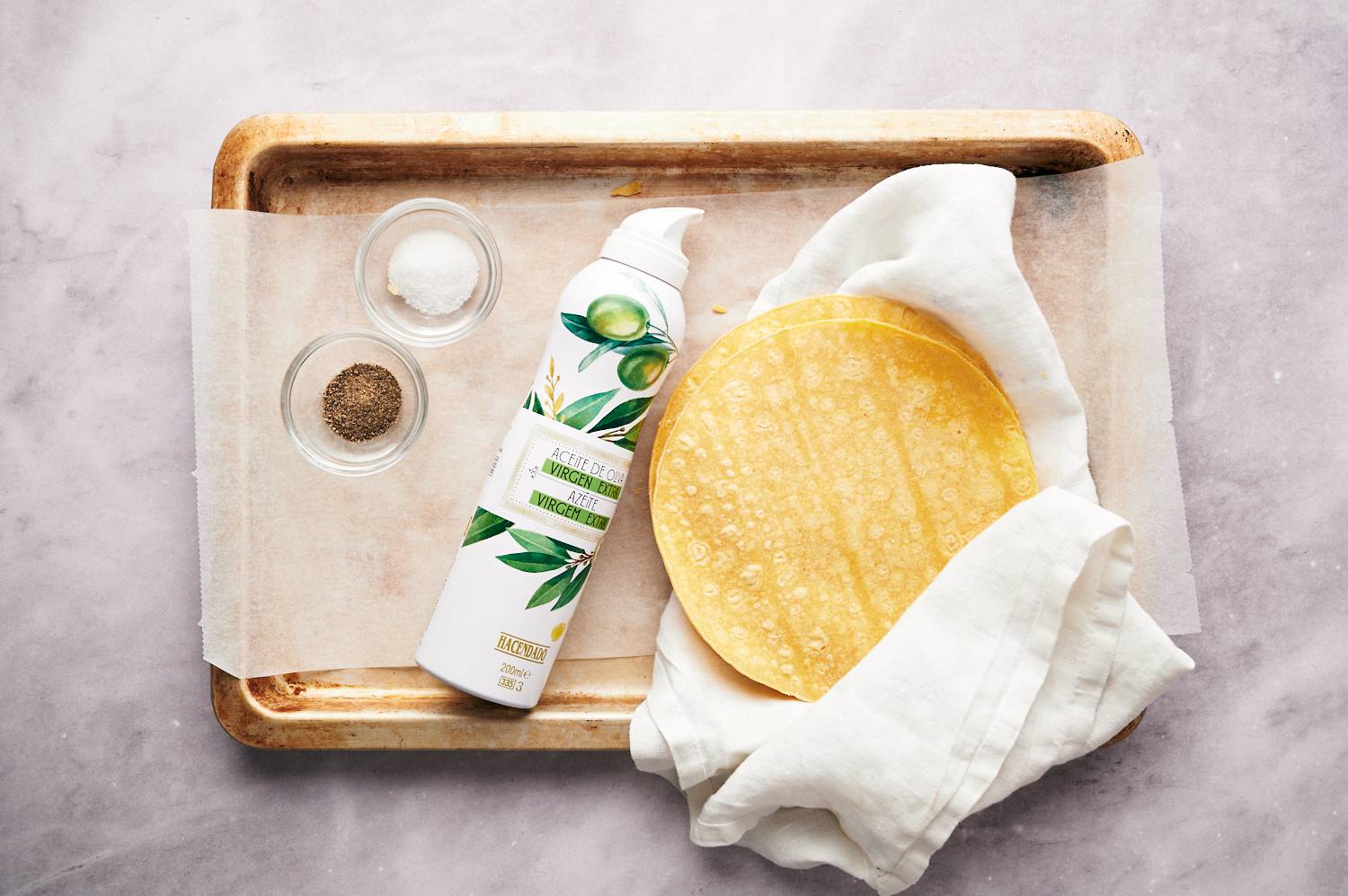 Ingredients for homemade baked tortilla chips including corn tortillas, cooking spray, and salt on a baking sheet.