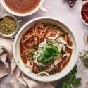 Birria de res with a red chile sauce served in a bowl with corn tortillas, sliced onions, cilantro, and consome.