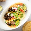 Black bean tacos with chipotle peppers served in corn tortillas topped with avocado, radish, tomatoes, and fresh cilantro.