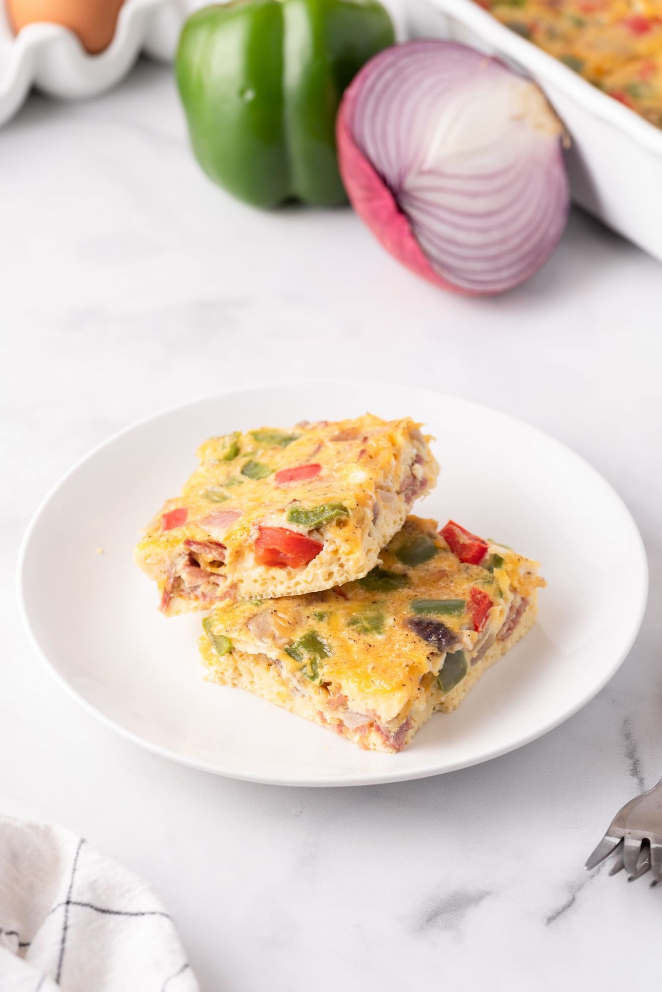 Two slices of egg casserole on a plate with peppers, turkey bacon, onion, and cheddar cheese. 