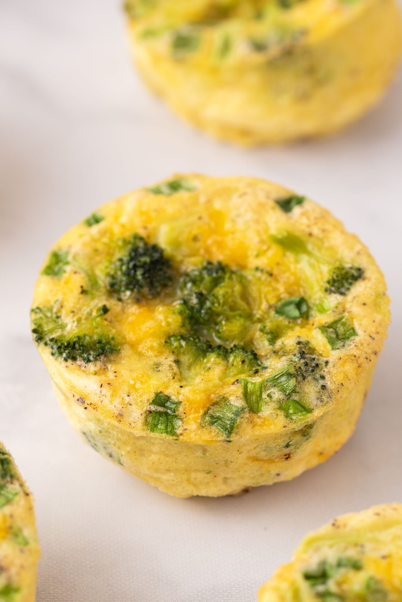 Egg muffin with broccoli pieces, green onions, and cheddar cheese on a white plate.