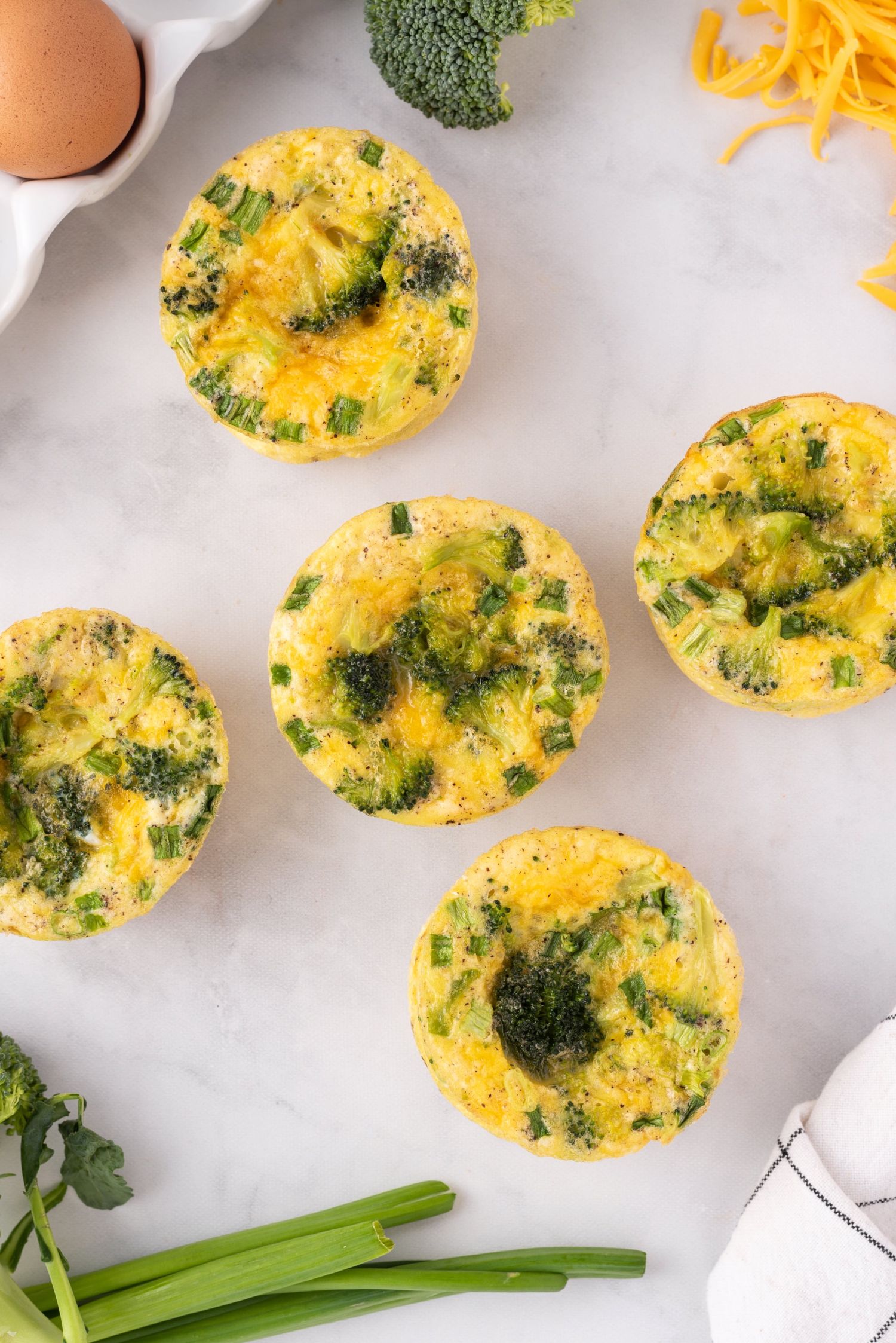 Mini egg, broccoli, and cheddar cheese frittatas on a white board with green onions.