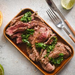 Carne asada marinated in citrus juices sliced thin on a plate with cilantro, limes, and orange slices.