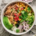 Carnitas burrito bowls with shredded pork, rice, black beans, corn, pico de gallo, lettuce, avocado, and lime wedges.