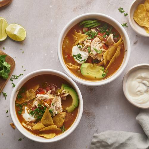 Slow Cooker Chicken Fajita Soup in tow bowls with shredded chicken, bell peppers, onions tortilla chips, and avocado.