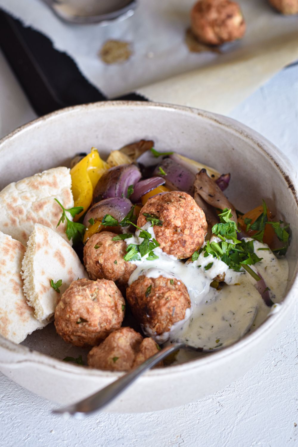 Ground chicken meatballs with feta cheese on a baking sheet and in a bowl with pita wedges, tzatziki, and vegetables.