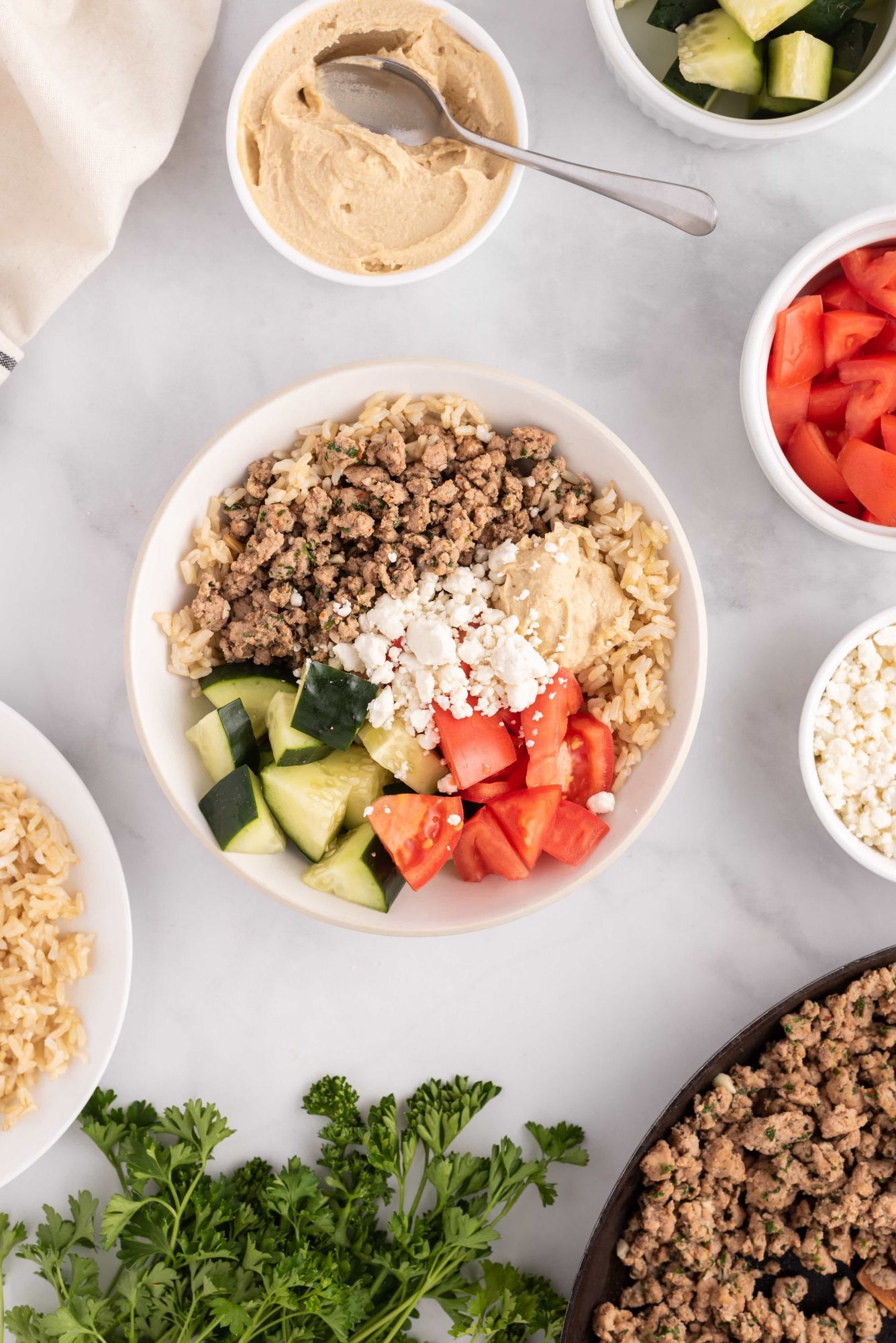 Ground chicken kofta in a bowl with cucumbers, tomatoes, brown rice, feta cheese, hummus, and parsley.