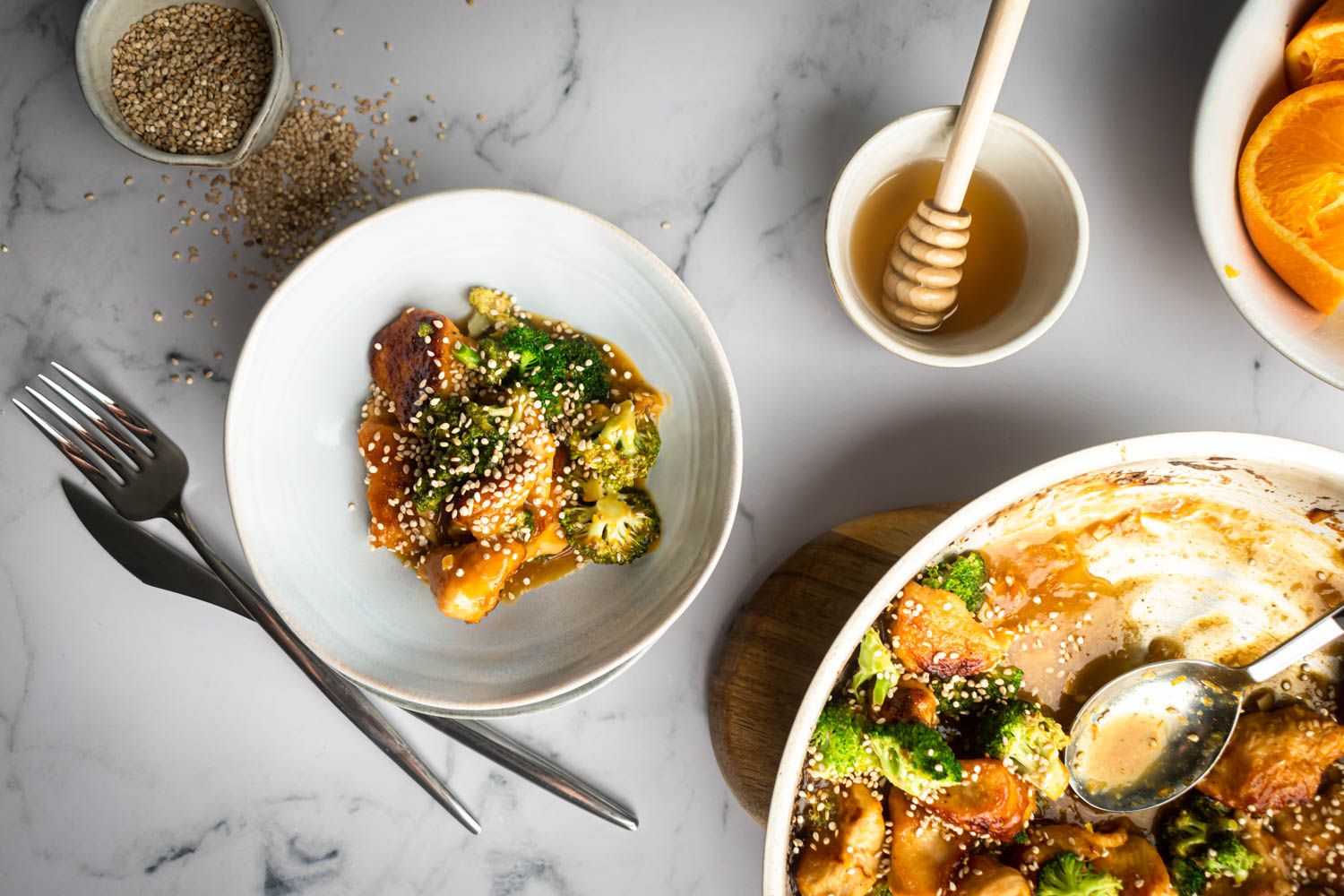 Orange chicken and broccoli served in a bowl with sesame seeds with honey and fresh oranges on the side.