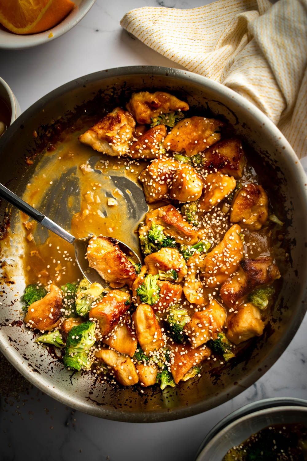 Baked orange chicken in a skillet with a thick, sticky sauce, broccoli, and sesame seeds.