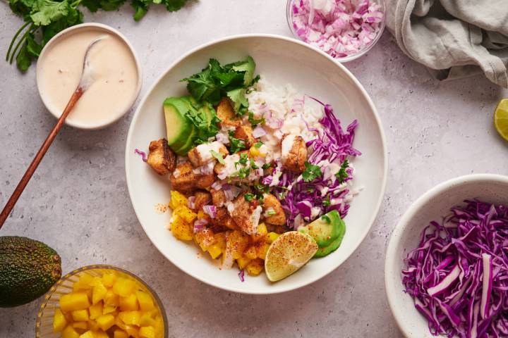 Fish taco bowls with flaky mahi mahi, avocado, mango, limes, shredded cabbage, cilantro, and taco sauce in a bowl with rice.