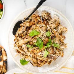 Greek chicken shredded on a plate with oregano, parsley, lemon, pita bread, and cucumber tomato salad.