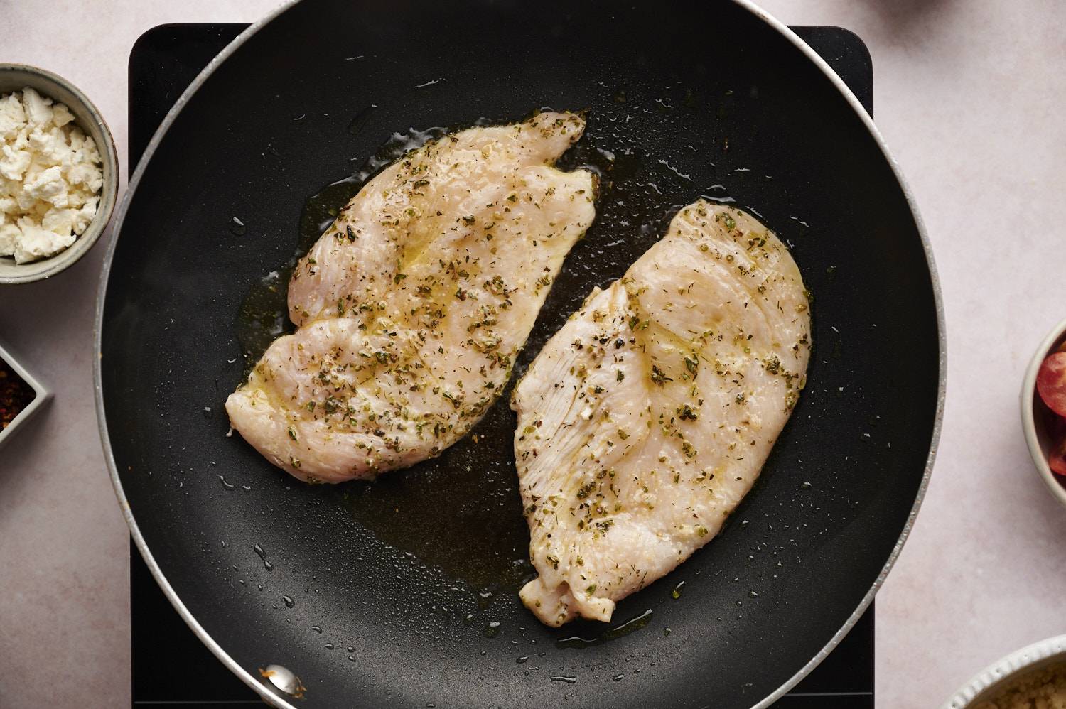 Greek chicken cooking in a skillet with oregano and dried dill for Greek chicken bowls.