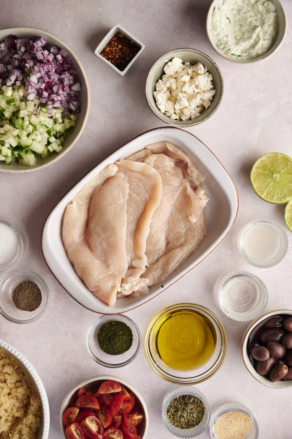 Ingredients for Greek Chicken Bowls including chicken breast, tomatoes, quinoa, cucumbers, olive oil, herbs, olives, and feta cheese.