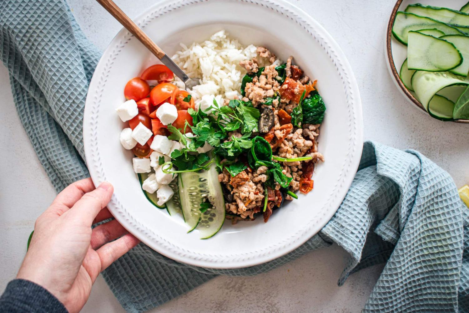 Greek turkey with rice, cherry tomatoes, olives, cucumbers, and ground turkey in a bowl with a blue napkin.