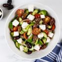 Greek salad with olives, feta cheese, cucumbers, tomatoes, and red onion in a white bowl with a blue napkin. 