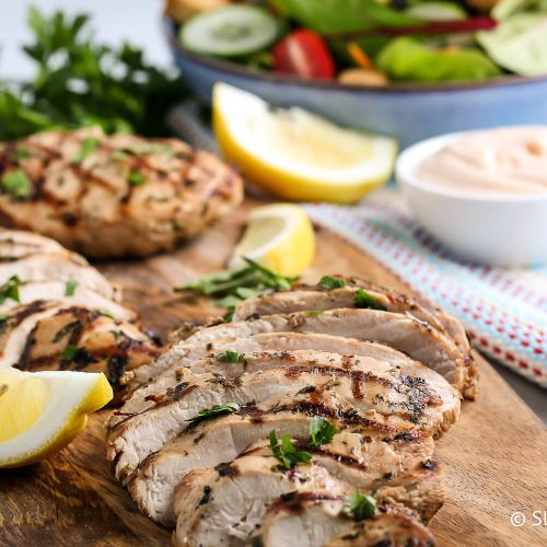 Grilled chicken breast sliced on a wooden cutting board garnished with herbs and lemon