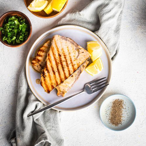 Grilled swordfish on a white plate seen from above with garlic, lemon, and herbs with fresh parsley.