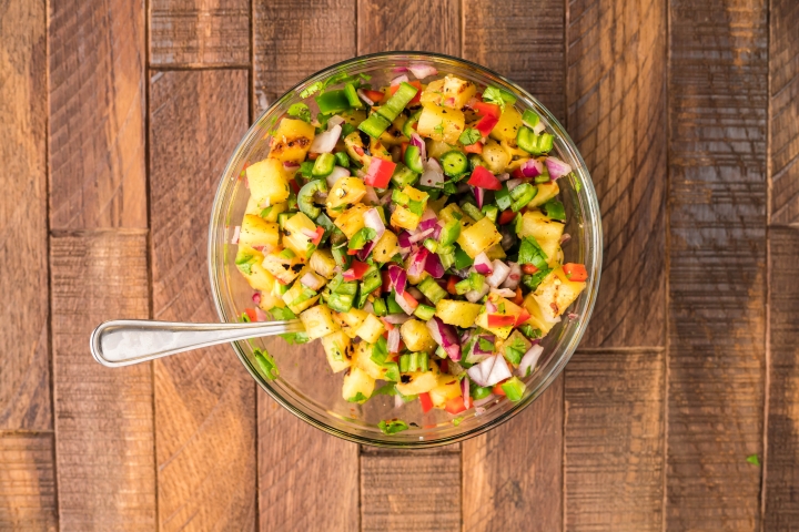 Grilled pineapple salsa served on steak tacos with chopped grilled pineapple, serrano peppers, red onion, red bell pepper, lime juice, and cilantro in a wooden bowl.