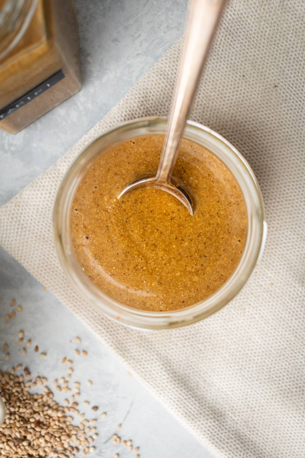 Creamy tahini made with sesame seeds in a glass jar with a spoon and extra sesame seeds on the side.