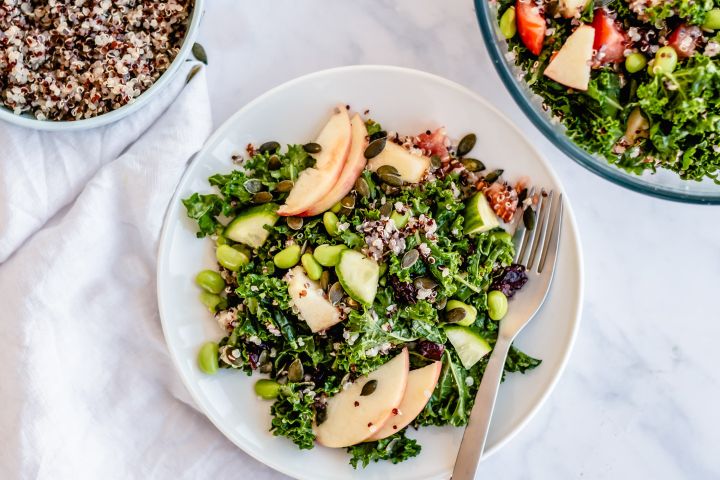 Honey lime kale and quinoa salad with sliced apples, dried cranberries, and pumpkin seeds in a bowl with cooked quinoa on the side.