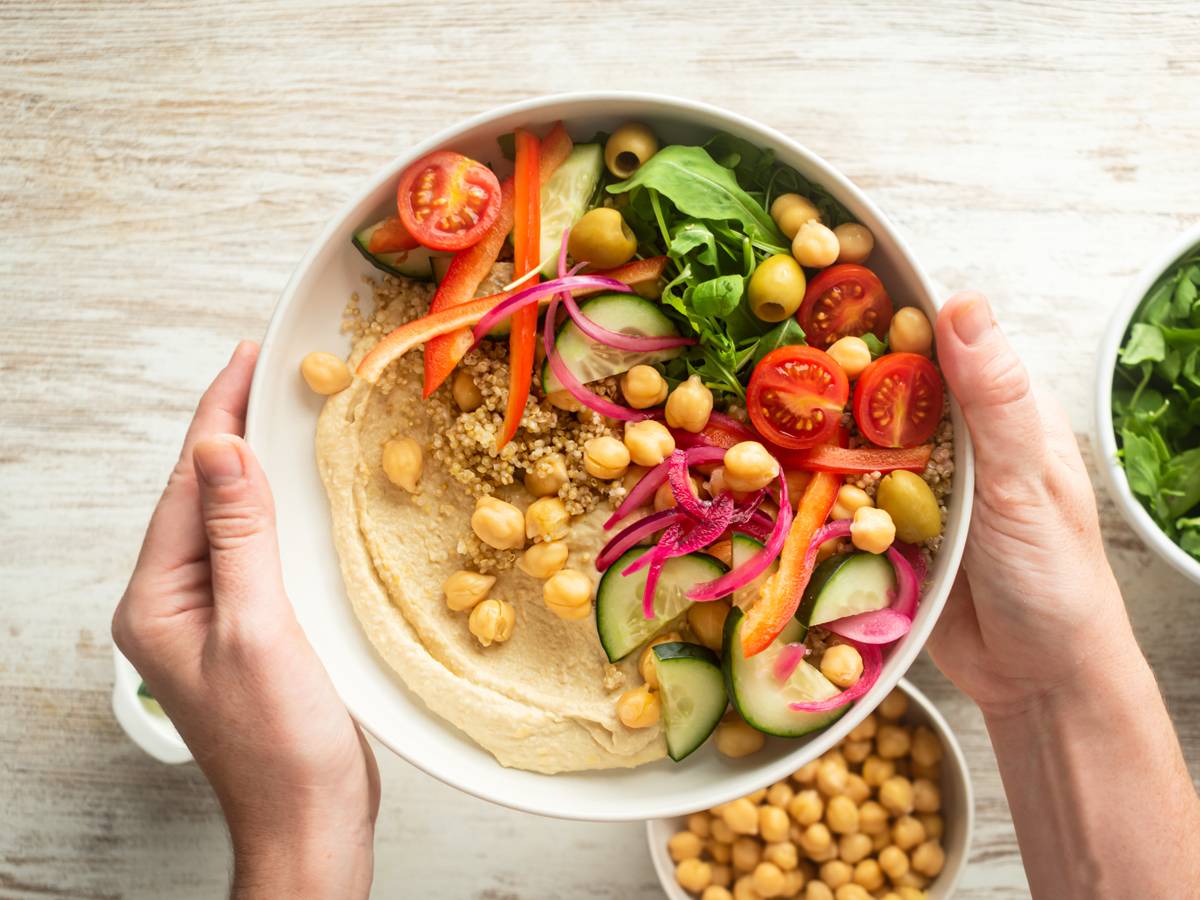 Woman hands holding a bowl with creamy hummus, quinoa, chickpeas, cucumbers, arugula, and red onions.