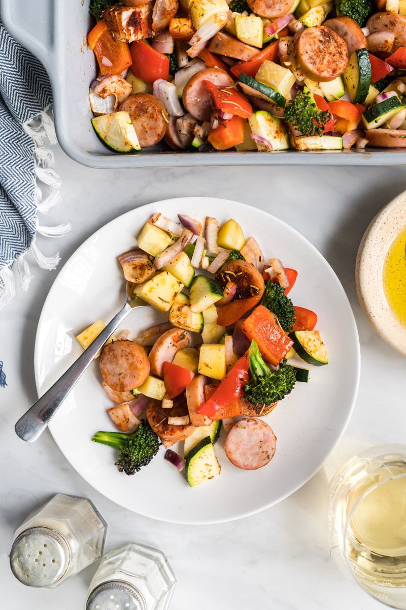 Baked sausage with potatoes, broccoli, onions, zucchini, and peppers in a baking dish and served on a plate.