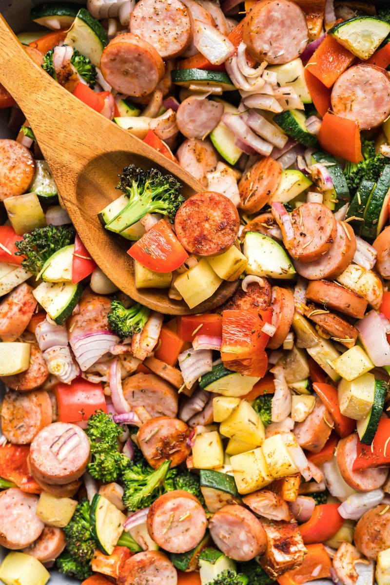 Sausage, broccoli, potatoes, and onions cooked in one baking dish with a wooden spoon.