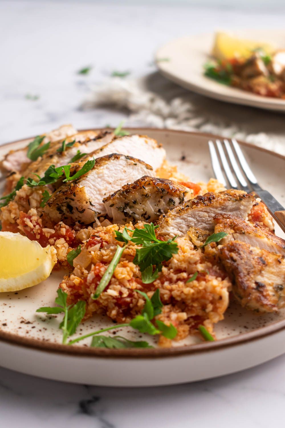 Sliced Parmesan chicken breast served over garlic cauliflower rice with tomatoes, parsley, and lemon,