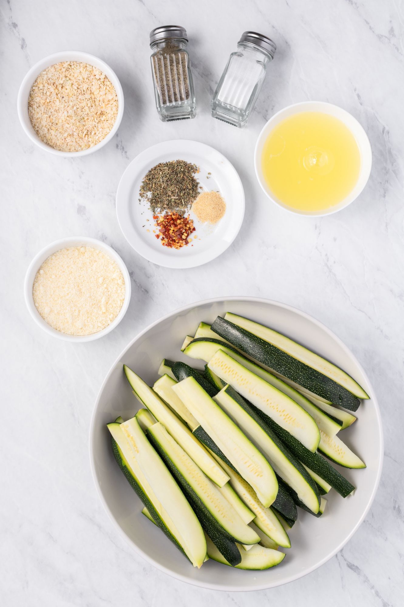 Ingredients for baked zucchini fries including sliced zucchini, eggs, breadcrumbs, and Parmesan cheese.