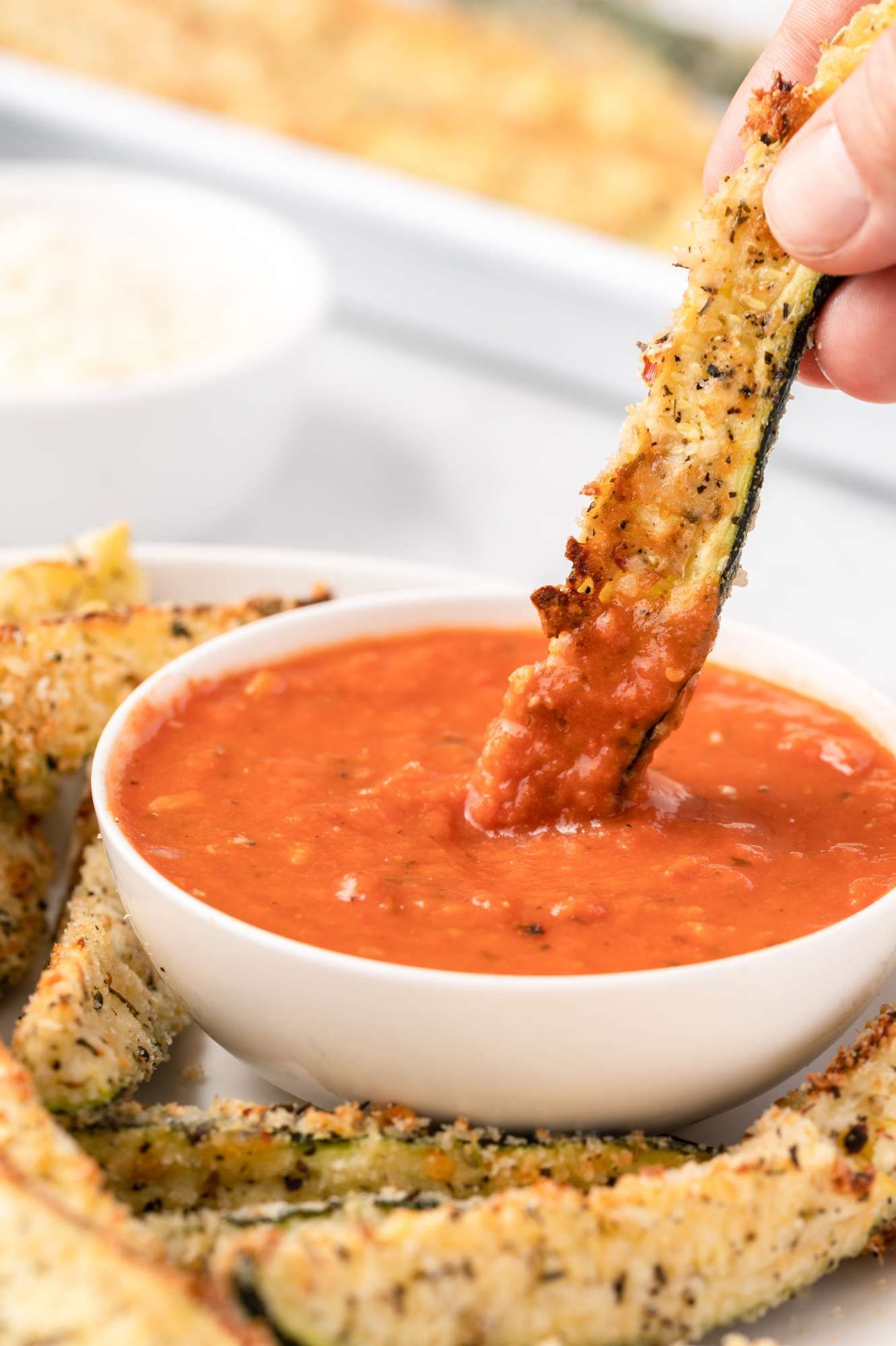 Baked zucchini fries being dipped in a bowl of marinara sauce with herbs.