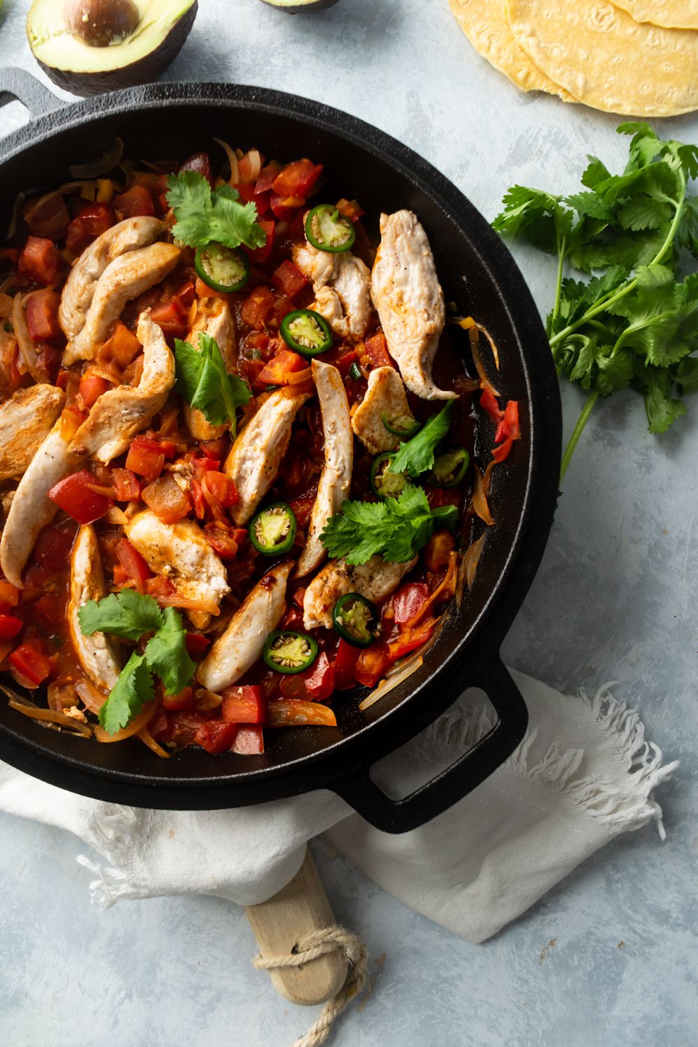 Chicken ranchero with tomatoes, onions, garlic, and peppers in a skillet with avocado and tortillas on the side.