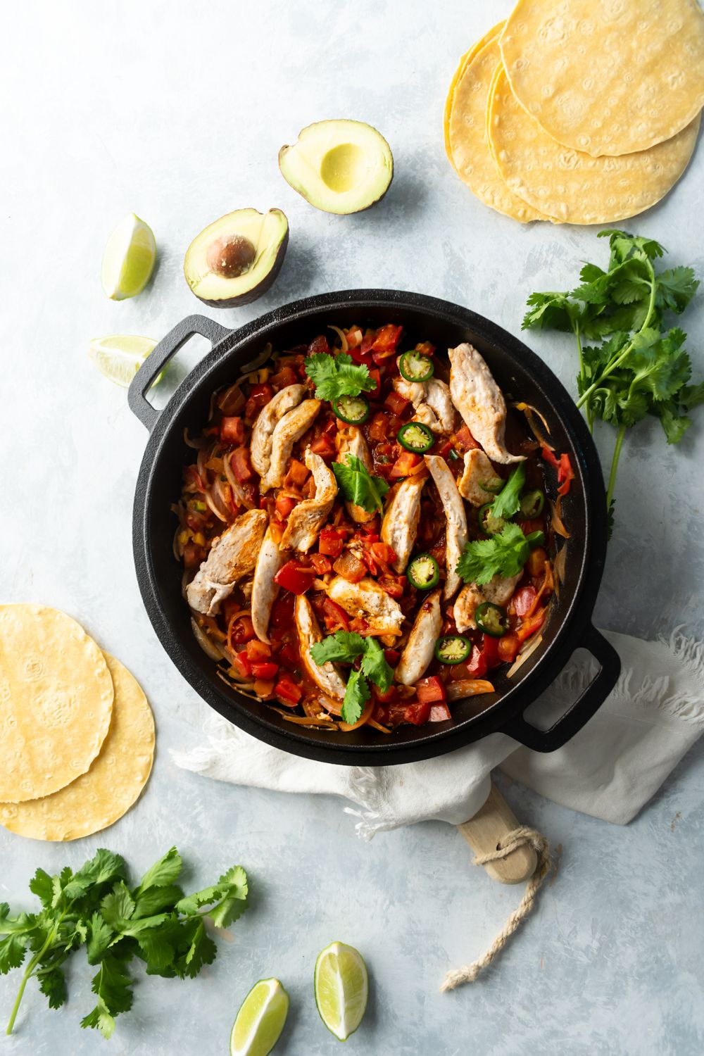 Chicken breast in ranchero sauce in a skillet with limes, cilantro, tortillas, and avocado.