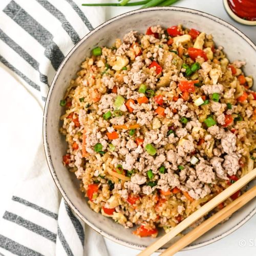 Cauliflower rice with ground pork, cabbage, carrots, and green onion in a bowl with chopsticks.