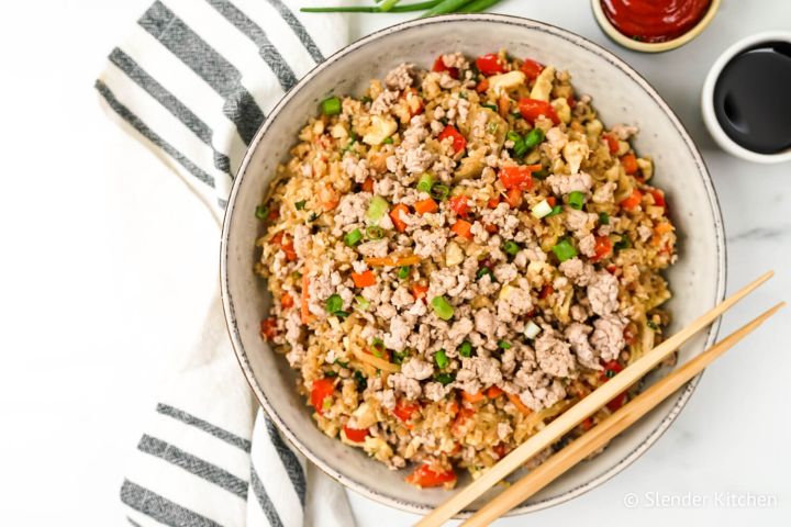 Cauliflower rice with ground pork, cabbage, carrots, and green onion in a bowl with chopsticks.