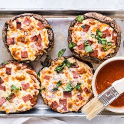 Portobello mushroom pizzas on a baking sheet with marinara sauce, melted cheese, basil, ham, and onions.