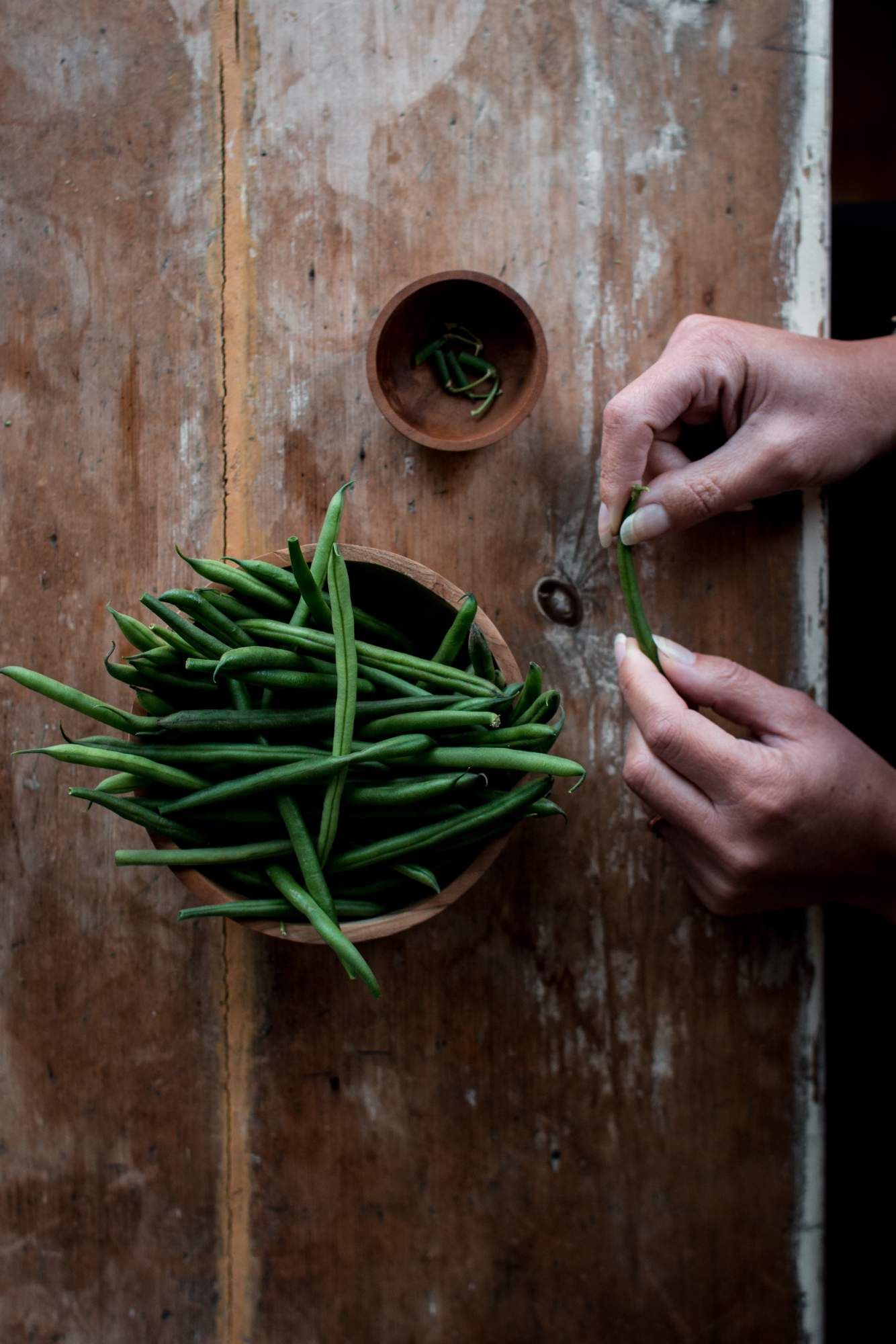 Green beans in a wooden bowl being prepped by a person removing the ends.