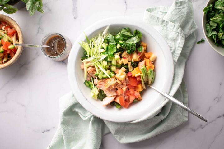 Salmon kale salad with sweet potatoes, avocado, apples, and a maple dijon dressing in a white bowl.