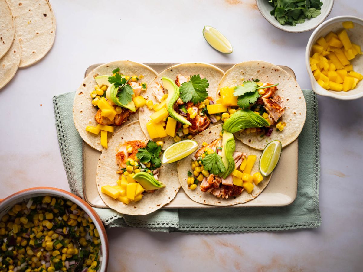 Salmon tacos with corn salsa, mango, fresh avocado slices, and lime wedges on a tray. 