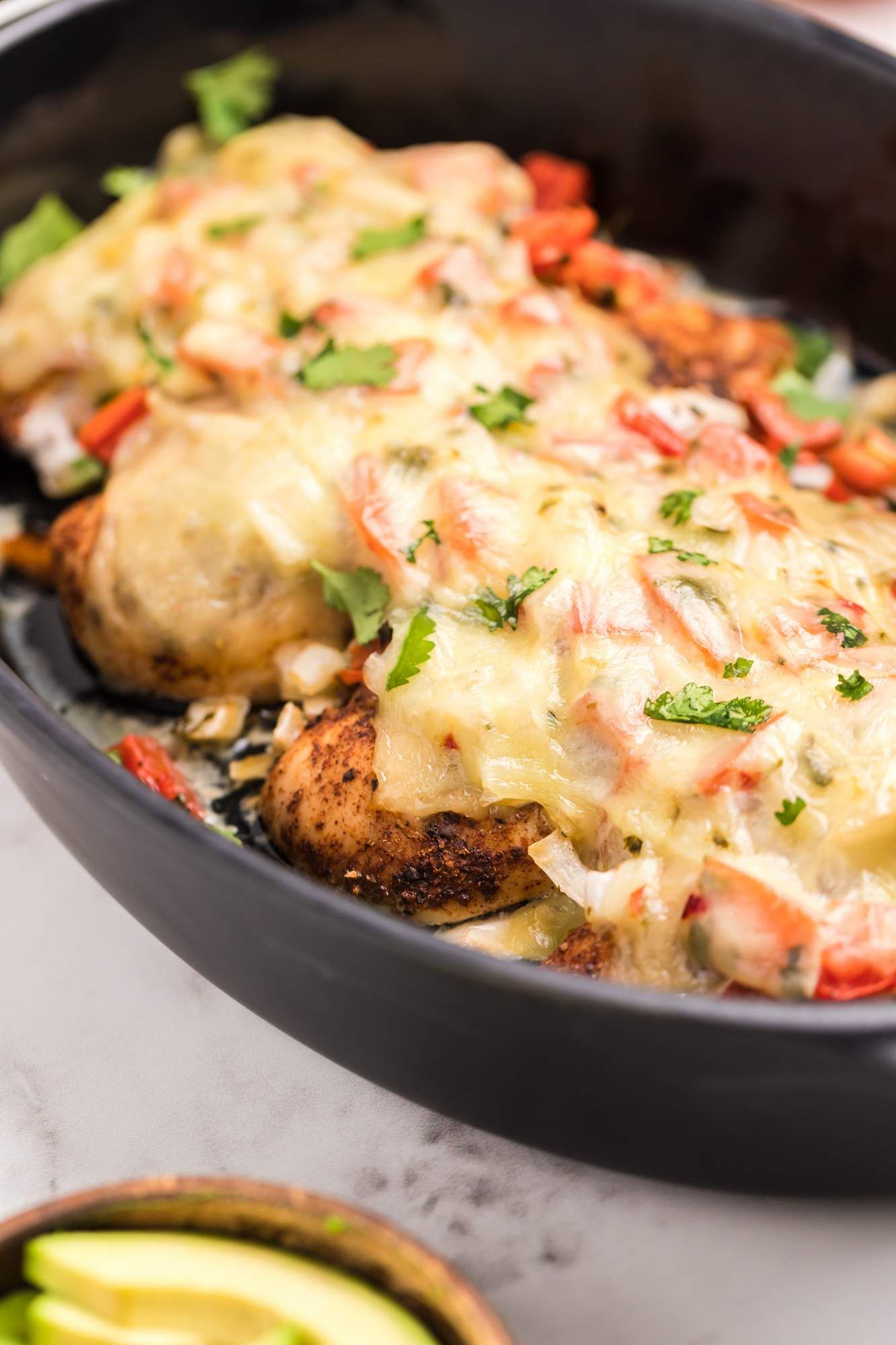 Baked chicken with pico de gallo salsa and melted cheese in a baking dish topped with avocado and cilantro.