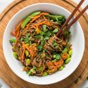 Asian noodle bowls with soba noodles, sesame dressing, carrots, edamame, and snap peas in a white bowl with chopsticks.