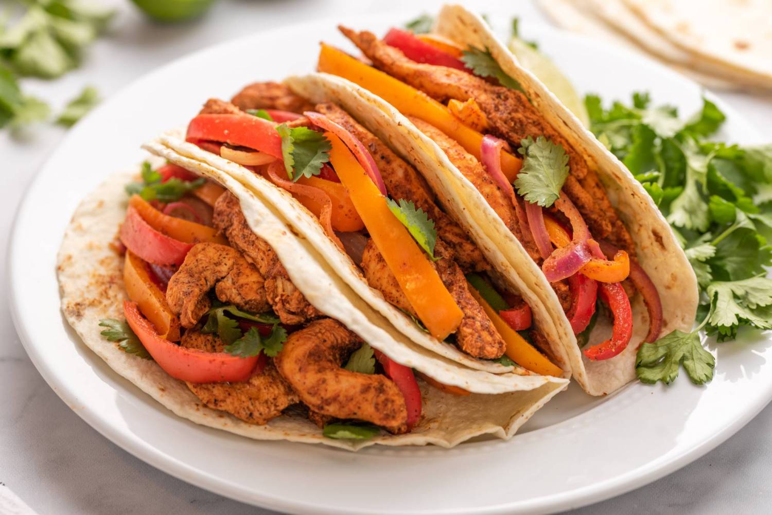 One pan chicken fajitas with red onions, bell peppers, and cilantro served in three flour tortillas on a plate.