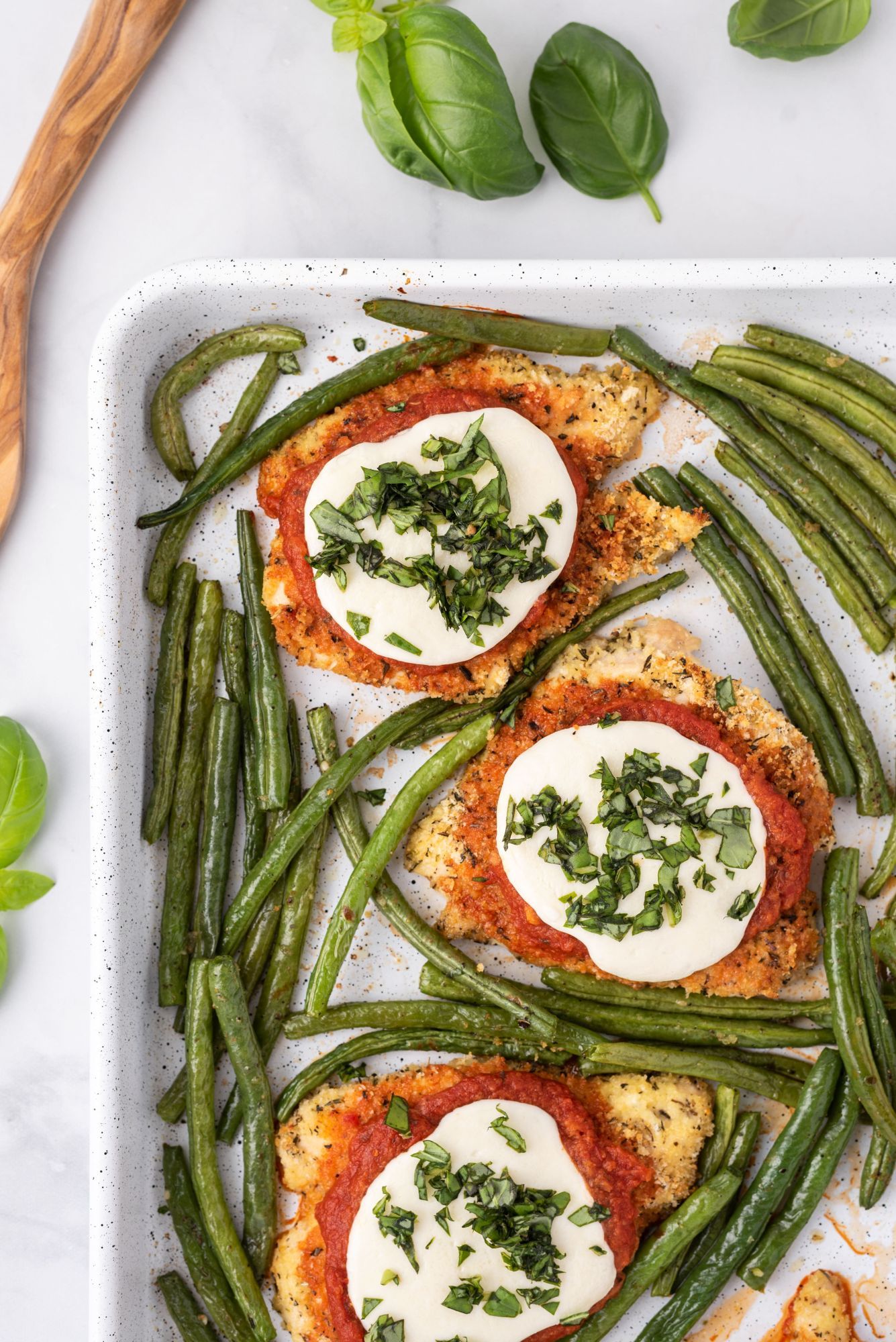 Baked chicken parmesan with marinara sauce, fresh mozzarella, basil, and green beans on a baking sheet.