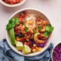 Shrimp taco bowls with cooked shrimp, avocado, rice, lime, cilantro, cabbage, corn, and pico de gallo in a white bowl.