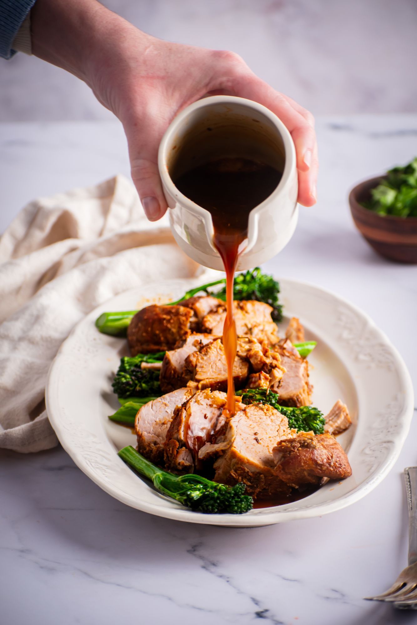 Sauce being drizzled on crockpot pork tenderloin on a plate with broccolini.