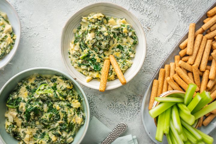 Slow cooker spinach artichoke dip with kale in three bowls with celery and breadsticks.